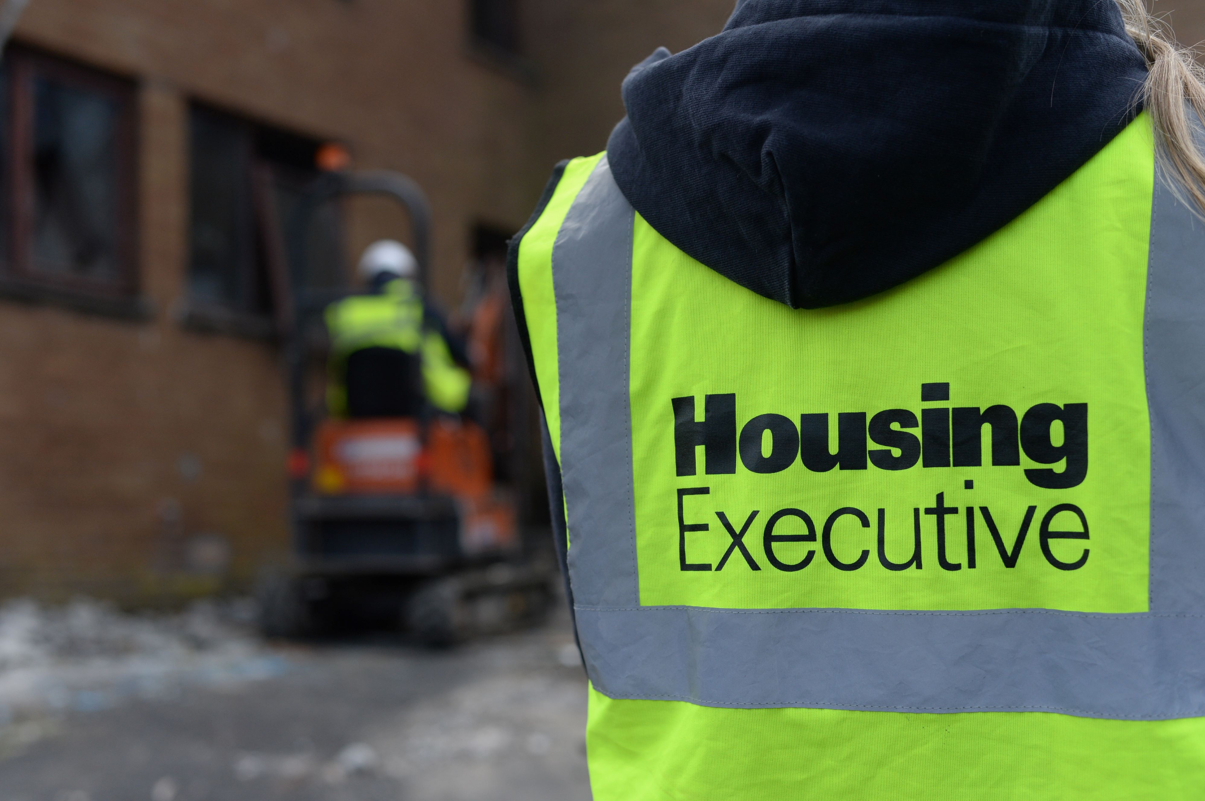 Contractor wearing high vis vest bearing words Housing Executive. Small earthmover at work in background near a red-brick house. 